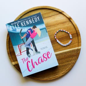 May include: A paperback book titled "The Chase" by Elle Kennedy, featuring a couple on an ice rink. The book cover is blue and pink with a hockey net and a hockey stick. A white beaded bracelet with the name "Fitzgerald" spelled out in black letters is on a wooden surface.