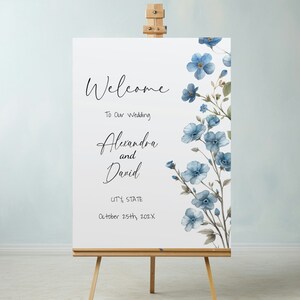 Pode incluir: Um cartaz de boas-vindas de casamento branco com flores azuis em aquarela. O cartaz diz "Bem-vindos ao nosso casamento Alexandra e David CIDADE, ESTADO 25 de outubro de 202X".