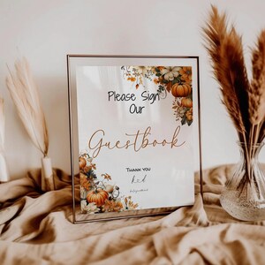 May include: A white sign with a gold frame and a floral design featuring pumpkins and leaves. The sign reads "Please Sign Our Guestbook" with the word "Guestbook" in a decorative script font. The sign also includes the text "Thank you" and a handwritten name.
