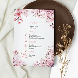 Może przedstawiać: Menu weselne z kwiatowym wzorem z różowymi kwiatami wiśni. Menu zawiera przystawki, dania główne, desery i napoje. Przystawki to sałatka sezonowa i ciasta krabowe. Dania główne to łosoś miodowy, stek wołowy z kością i indyk pieczony w ziołach. Desery to tarta owocowa, lody waniliowe z brownie. Napoje to napoje bezalkoholowe i woda.