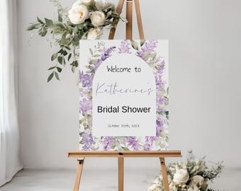 May include: A white welcome sign with purple flowers and greenery around the border. The sign says "Welcome to Katherine's Bridal Shower, October 25th, 202X".