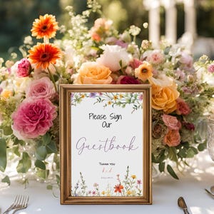 Pode incluir: Um sinal com moldura dourada com uma borda floral e o texto "Please Sign Our Guestbook" em escrita cursiva roxa. O sinal está sobre uma mesa com um centro de mesa floral.