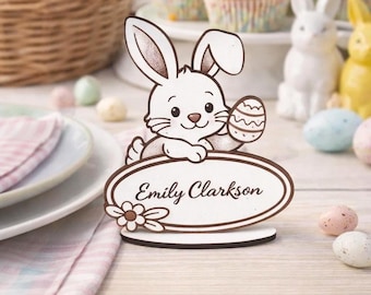 Op de afbeelding: Een witte paashaas-naamkaarthouder met de naam "Emily Clarkson" op een ovaal bord. Het konijn houdt een versierd paasei vast. Andere paasdecoraties zijn cupcakes, eieren en konijnenbeeldjes.