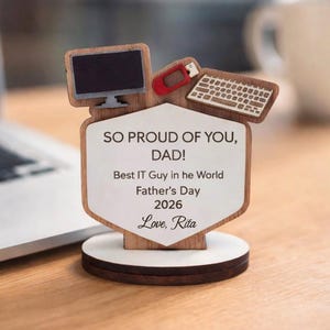 Op de afbeelding: Een houten Vaderdag bureau-ornament met een computerthema. Het heeft een monitor, toetsenbord en USB-stick. De tekst luidt: "SO PROUD OF YOU, DAD! Best IT Guy in he World Father's Day 2026 Love, Rita."