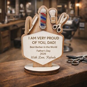 Könnte beinhalten: Ein hölzernes Vatertagsgeschenk für einen Friseur. Die Tafel zeigt Friseurwerkzeuge, eine Barbierstange und den Text "I AM VERY PROUD OF YOU, DAD!". Es enthält den Text "Best Barber in the World", "Father's Day 2026" und "With Love, Rafaela".