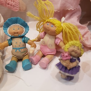 May include: A collection of Cabbage Patch Kids dolls, including a doll wearing a blue and white polka dot dress, a doll wearing a pink dress, and a doll wearing a purple dress. The dolls are all small and have blonde hair.
