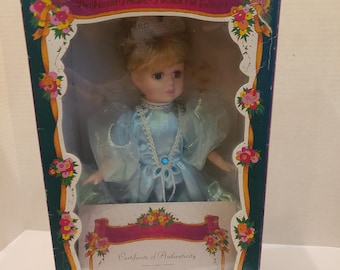 Puede incluir: Una muñeca de porcelana con un vestido azul claro, una corona y una gema azul en el pecho. La muñeca está en una caja con un certificado de autenticidad y el texto "Fairytale Treasures" y "Limited Collectors Edition".