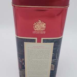 K&ouml;nnte beinhalten: Rote Blechdose mit einer Vintage-Illustration einer Pferdekutsche und Personen vor einem Geb&auml;ude. Der Text auf der Dose lautet "Johnnie Walker: Der Mann, die Legende, der Whisky."