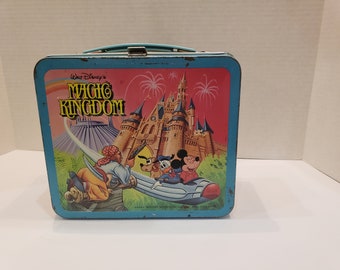 May include: Vintage metal lunchbox with a blue frame and handle. The front features the text "Walt Disney's MAGIC KINGDOM" above an illustration of a castle, fireworks, and Disney characters. The lunchbox has some wear and tear.