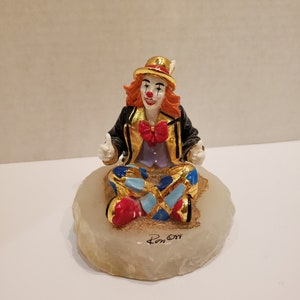 May include: A small, hand-painted porcelain figurine of a clown sitting cross-legged on a white marble base. The clown is wearing a black jacket with gold trim, a red bow tie, and a blue and white checkered pattern on their pants. The clown has red hair, a white face with red painted lips and cheeks, and a black hat with a feather.