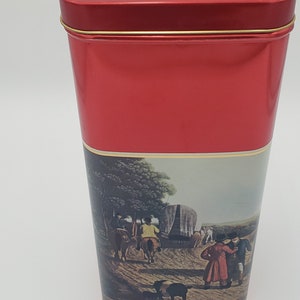 K&ouml;nnte beinhalten: Rote Metalldose mit goldener Einfassung. Die Dose zeigt eine Vintage-Illustration von Menschen, die in einem von Pferden gezogenen Wagen reisen. Der Text "Johnnie Walker" ist auf der Oberseite der Dose aufgedruckt.