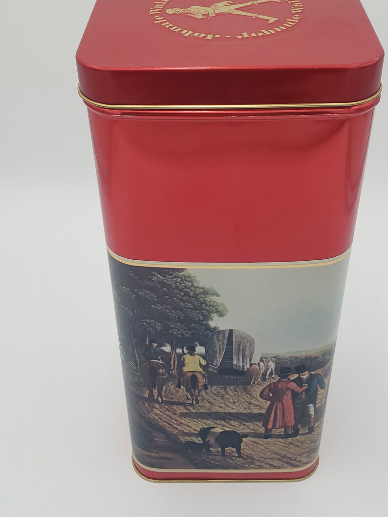 K&ouml;nnte beinhalten: Rote Metalldose mit goldener Einfassung. Die Dose zeigt eine Vintage-Illustration von Menschen, die in einem von Pferden gezogenen Wagen reisen. Der Text "Johnnie Walker" ist auf der Oberseite der Dose aufgedruckt.