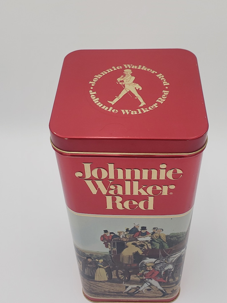 K&ouml;nnte beinhalten: Rote Blechdose mit einem Johnnie Walker Red-Etikett und einer Vintage-Illustration einer Kutsche, die von Pferden gezogen wird. Die Oberseite der Dose hat ein kreisf&ouml;rmiges Logo mit dem Text "Johnnie Walker Red" und einem Mann, der mit einem Stock geht.