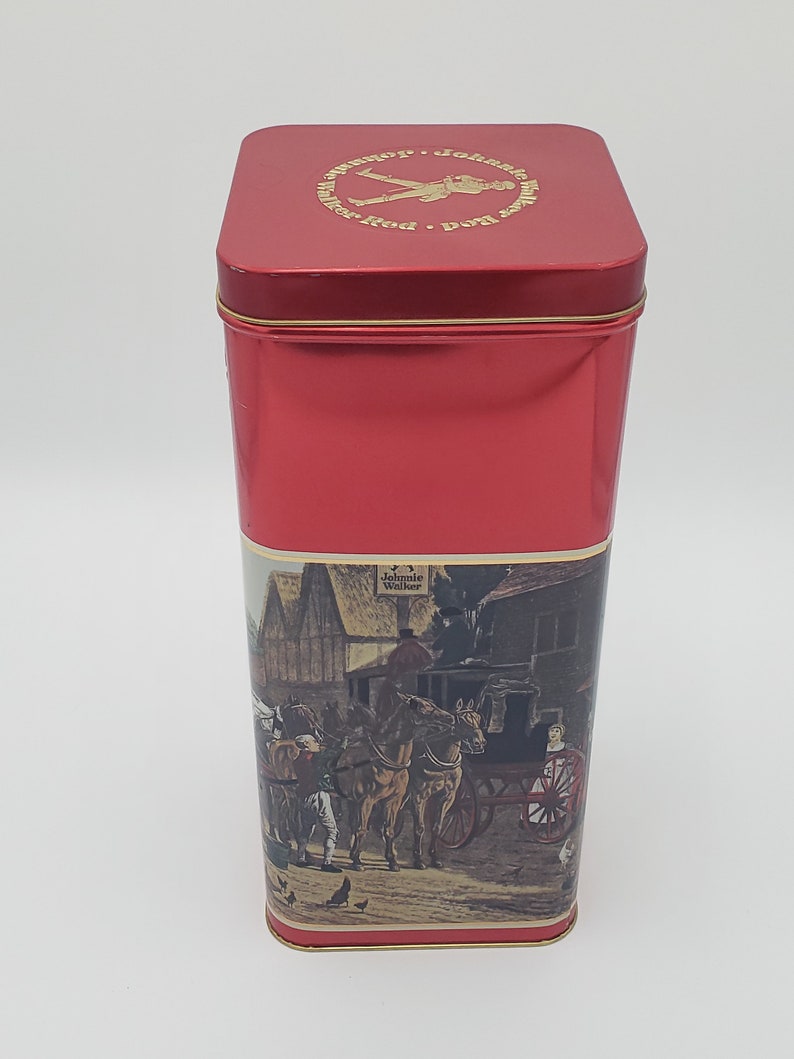 K&ouml;nnte beinhalten: Rote Metalldose mit einem goldenen Etikett, auf dem ein Mann zu sehen ist, der geht, und der Text "Johnnie Walker Red Label". Das Etikett zeigt auch eine Szene mit einer von Pferden gezogenen Kutsche mit Personen vor einem Geb&auml;ude.