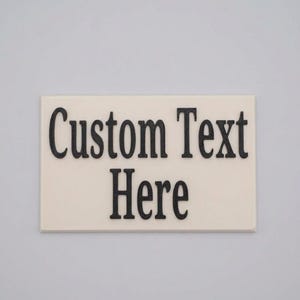 Könnte beinhalten: Ein rechteckiges, cremeweißes Schild mit dem schwarzen, erhabenen Text Custom Text Here. Die Beschriftung ist in einer fetten Serifenschrift gehalten und bildet einen klaren Kontrast zur hellen, glatten und flachen rechteckigen Grundfläche.