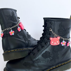 Puede incluir: Botas de cuero negro con bordado floral y cadenas plateadas con dijes rosas y azules. Los dijes incluyen una cara de gato y flores.
