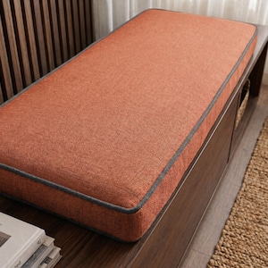 May include: A rectangular bench cushion featuring a textured, burnt orange fabric surface with dark grey piping along the edges. The cushion is designed with a firm, flat top and straight sides, showcasing a consistent weave pattern throughout.