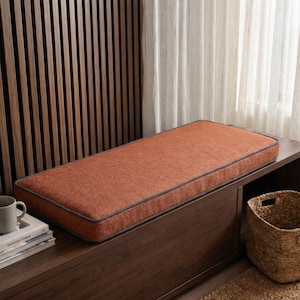 May include: A rectangular bench cushion featuring a textured, burnt orange fabric surface with dark grey piping along the edges. The cushion is designed with a flat, firm profile and a consistent weave pattern across the top and side panels.