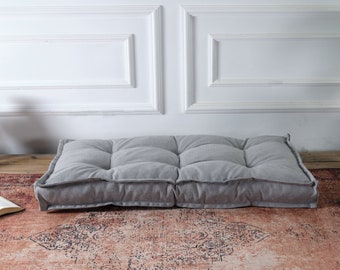 Puede incluir: Un cojín de suelo rectangular de tela gris claro con un diseño capitoné y costuras visibles. El cojín tiene una construcción gruesa y acolchada con bordes elevados y una superficie texturizada, ofreciendo una opción de asiento plana.