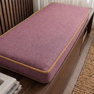 May include: A rectangular bench cushion featuring a textured plum-coloured fabric top and sides, accented with a thick mustard-yellow piping trim along the edges. The cushion is positioned on a dark wooden base with a visible storage compartment.