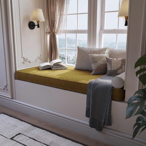 Puede incluir: Un asiento de ventana con un cojín de color mostaza, adornado con varias almohadas en tonos neutros. También hay una manta con estampado gris y blanco y un libro abierto. La escena está iluminada por luz natural y apliques de pared.