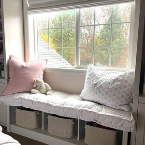 May include: A white window seat bench featuring three open storage cubbies with woven beige baskets. The bench is topped with a long, tufted white cushion, a solid light pink square pillow, a white patterned quilted pillow, and a small plush toy.