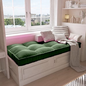 May include: A cozy window seat with a tufted, dark green cushion and a white wooden base. Decorative pillows in various patterns and colors are arranged on the seat, along with a folded blanket. The window offers a view of a cityscape.