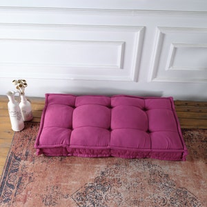 May include: A rectangular, tufted floor cushion in a vibrant magenta color. The cushion features a grid of stitched indentations across its surface, creating a plush, segmented appearance with raised edges and a thick, soft construction.