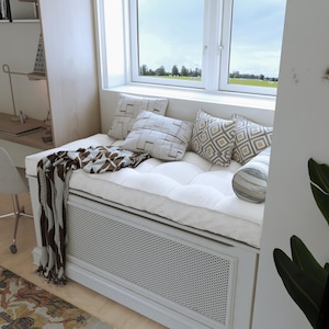 May include: A white window seat with a white cushion and a brown and white patterned throw blanket. The seat has a white, gray, and brown patterned pillow, a gray and white patterned pillow, and a beige and brown patterned pillow. The seat is in front of a window with a view of a green field.