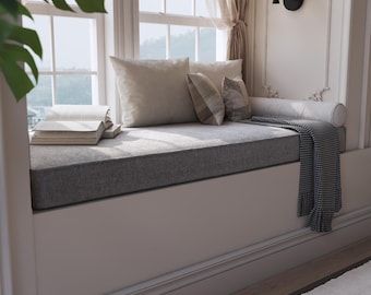 May include: A comfortable window seat featuring a grey cushion and neutral-toned pillows. A striped blanket and a stack of books enhance the inviting feel. The window is framed with white trim and sheer curtains.
