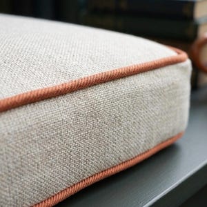 May include: Close-up of a square, beige cushion with a textured fabric. The cushion has a thin, orange-brown trim around the edges. The fabric appears to be a linen or similar material, with a subtle, natural texture.