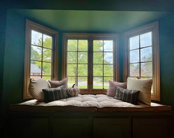 May include: A bay window seat featuring a tufted beige cushion, accented with several decorative pillows in striped and solid patterns, and a small fabric pumpkin. The window frame is made of wood, set against dark green walls.