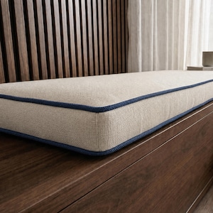 May include: A rectangular bench cushion featuring a textured beige fabric surface with a distinct dark blue piping trim along the edges. The cushion is designed with a firm, flat profile and a clean, structured finish for a seating area.