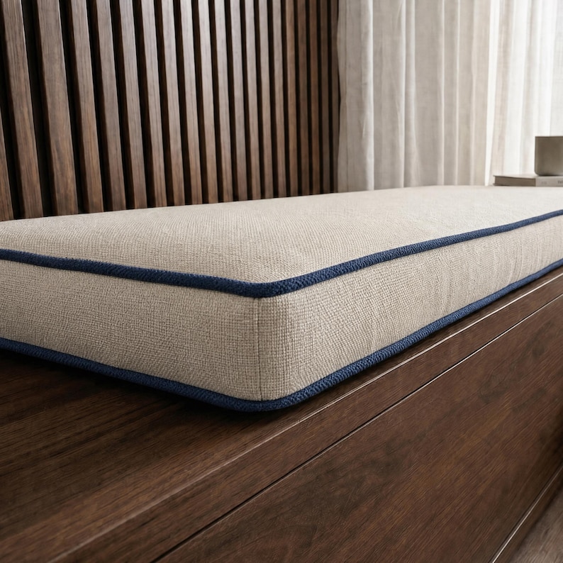 May include: A rectangular bench cushion featuring a textured beige fabric surface with a distinct dark blue piping trim along the edges. The cushion is designed with a firm, flat profile and a clean, structured finish for a seating area.