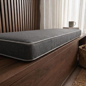 May include: A rectangular bench cushion upholstered in a dark gray textured fabric with light gray piping along the edges. The cushion is positioned on a flat, smooth, dark wood surface, showcasing a clean, structured, and firm rectangular shape.