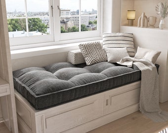 May include: A charcoal grey tufted bench cushion sits atop a white wooden storage bench with cabinet doors. The cushion is accented by several decorative throw pillows in neutral tones of white, grey, and black, along with a draped herringbone throw.
