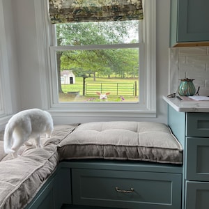 May include: A white cat stands on a beige tufted bench cushion next to a window. Outside, a white rabbit with long ears is visible near a green metal gate and a small white building. The window frame is white, and the cabinetry is blue-green.