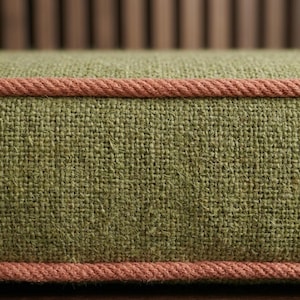 May include: A rectangular cushion featuring a coarse, olive green woven fabric texture. The cushion is bordered by a thick, twisted rope-style trim in a muted salmon pink color that runs horizontally along the top and bottom edges of the piece.