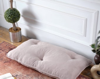 Puede incluir: Un cojín de suelo rectangular, color rubor, con mechones de botones. El cojín está colocado sobre una alfombra estampada, cerca de una pared blanca y un topiario en maceta. Diseñado para sentarse o descansar en el suelo.