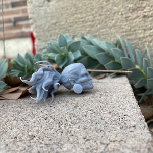 Puede incluir: Una figurita gris de pl&aacute;stico impresa en 3D que representa una cabeza humana con cabello largo. La figurita est&aacute; sentada sobre una superficie de concreto gris.