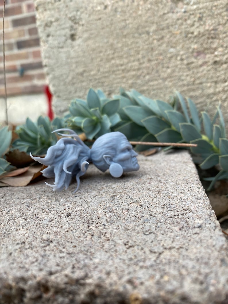 Puede incluir: Una figurita gris de pl&aacute;stico impresa en 3D que representa una cabeza humana con cabello largo. La figurita est&aacute; sentada sobre una superficie de concreto gris.
