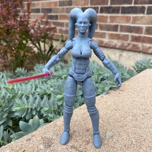 Puede incluir: Figura de acción gris de un personaje alienígena femenino, sosteniendo un sable de luz rojo. La figura tiene una armadura detallada y colas de cabeza distintivas. La figura está de pie sobre una superficie de hormigón con vegetación de fondo.