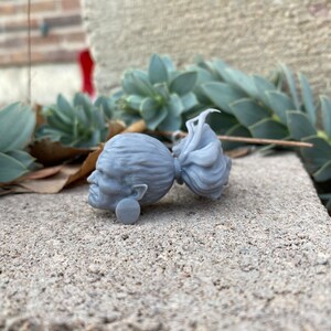 Puede incluir: Una figurita de cabeza de mujer con una trenza larga impresa en 3D de color gris. La figurita est&aacute; tumbada de lado sobre una superficie de hormig&oacute;n gris.