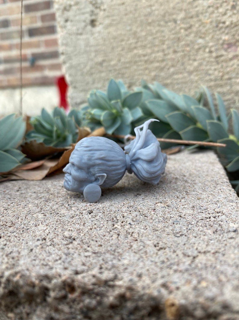 Puede incluir: Una figurita de cabeza de mujer con una trenza larga impresa en 3D de color gris. La figurita est&aacute; tumbada de lado sobre una superficie de hormig&oacute;n gris.