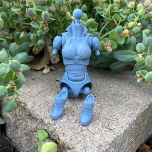 May include: A light blue, headless action figure torso with detached arms and legs. The figure has a high collar, button details, and a sculpted bodice. The detached legs include a pair of matching blue boots. The figure is made of plastic.
