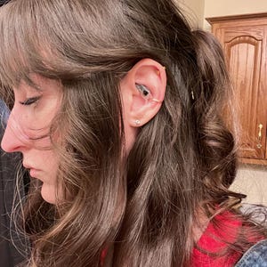 May include: A woman with long brown hair wearing a red shirt and blue overalls. She has a gold hoop earring in her upper ear and a small stud earring in her lower ear.