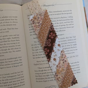 May include: A fabric bookmark featuring a diagonal patchwork design of various brown, white, and floral patterned fabrics with scalloped edges. The bookmark is topped with a piece of cream-colored lace and is placed inside an open book.