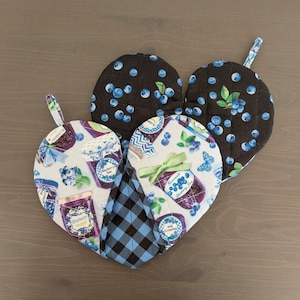 May include: Heart-shaped quilted oven mitts and pot holders. The mitts feature a black background with blue blueberries, and a light blue background with blueberry jam jars. A blue and black plaid pot holder is also included.