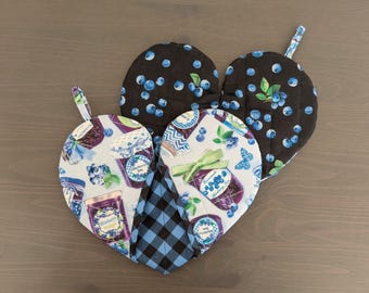 May include: Heart-shaped quilted oven mitts and pot holders. The mitts feature a black background with blue blueberries, and a light blue background with blueberry jam jars. A blue and black plaid pot holder is also included.