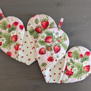 May include: Two pink and white heart-shaped pot holders with a strawberry print. The pot holders have a quilted texture and are perfect for protecting your hands while cooking.